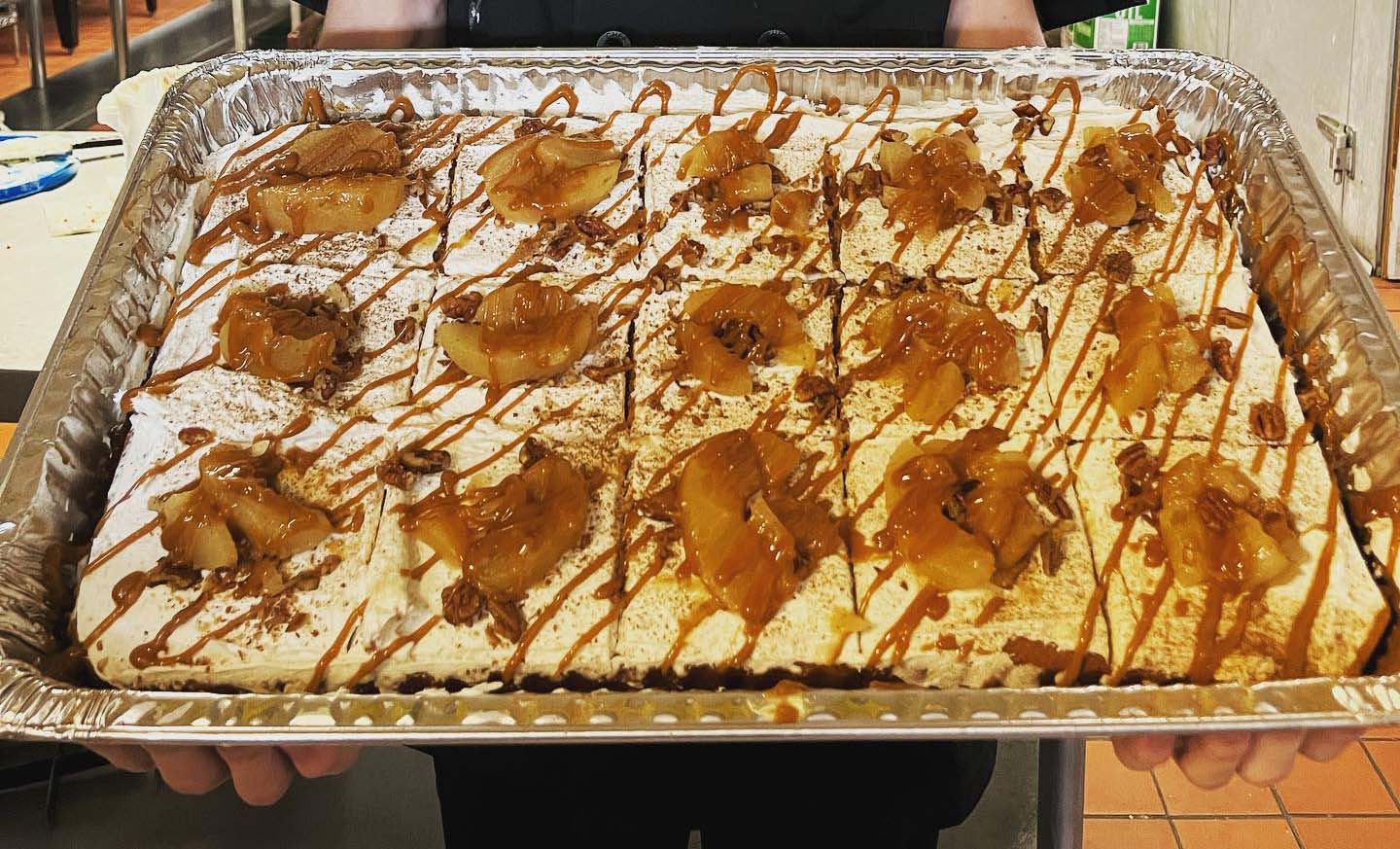 Large honey bun cake in a foil pan topped with icing, caramel drizzle, and apple slices