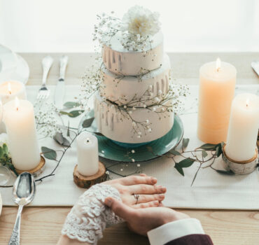 Wedding Cake Ideas You’ll Love — Why a Honey Bun Cake Steals the Show A partial view of newlyweds holding hands while seated at a styled wedding table featuring a rustic tiered wedding cake, candles, and greenery accents. Photo by KateNovikova, licensed via Depositphotos.