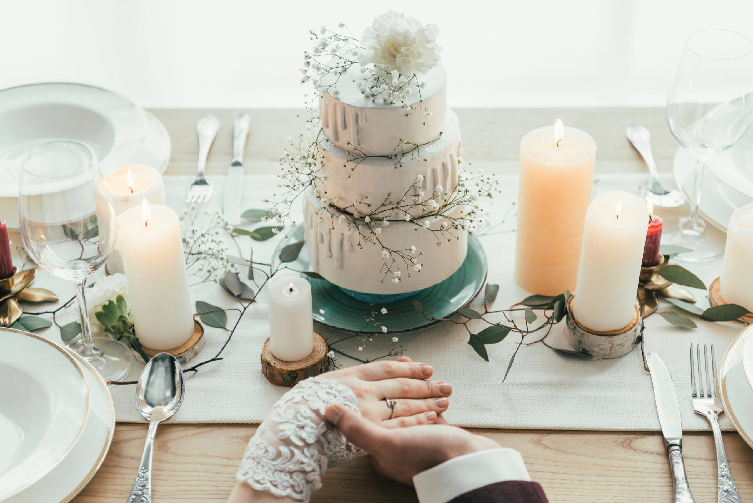 Wedding Cake Ideas You’ll Love — Why a Honey Bun Cake Steals the Show A partial view of newlyweds holding hands while seated at a styled wedding table featuring a rustic tiered wedding cake, candles, and greenery accents. Photo by KateNovikova, licensed via Depositphotos.
