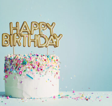 Why a Honey Bun Cake Is the Ultimate Birthday Cake for Any Celebration in Bryans Road A white frosted birthday cake decorated with colorful sprinkles and a gold “Happy Birthday” topper, photographed against a soft blue background. Photo by RuthBlack, licensed via Depositphotos.