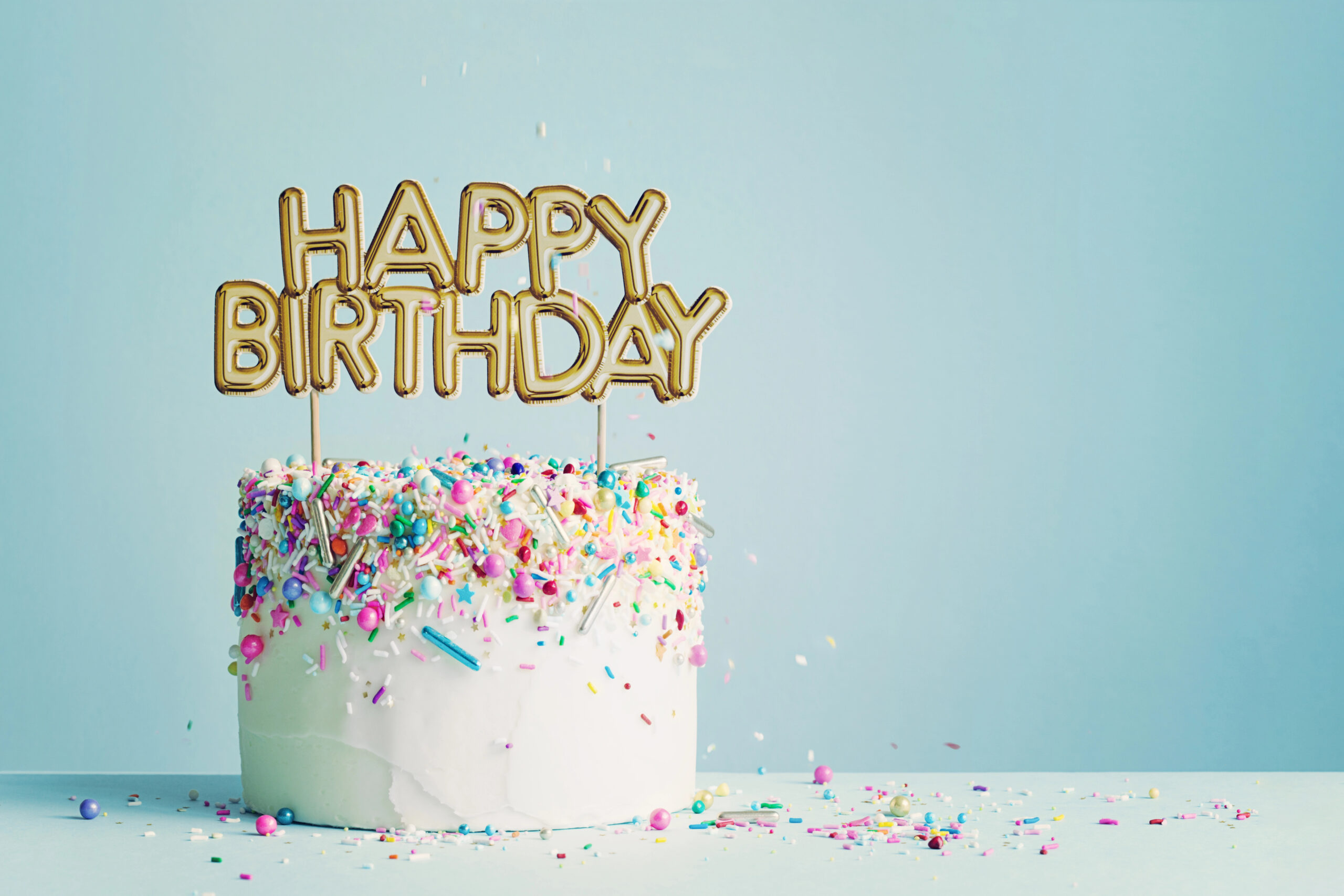 Why a Honey Bun Cake Is the Ultimate Birthday Cake for Any Celebration in Bryans Road A white frosted birthday cake decorated with colorful sprinkles and a gold “Happy Birthday” topper, photographed against a soft blue background. Photo by RuthBlack, licensed via Depositphotos.