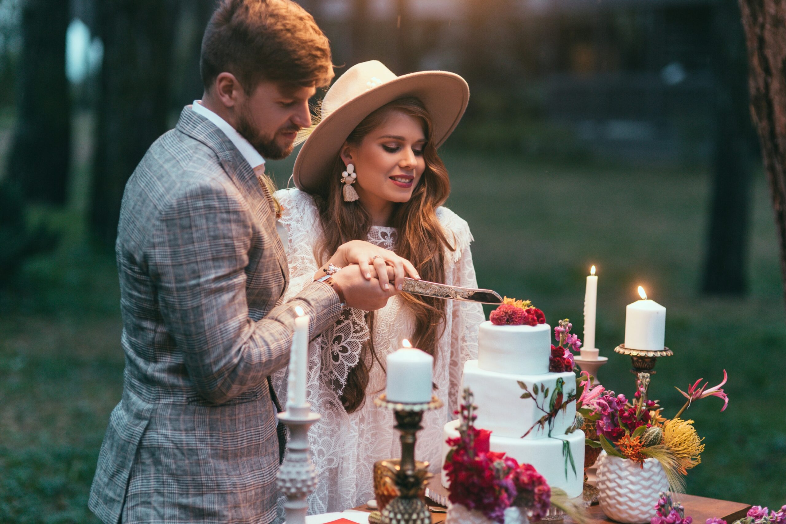 A bride and groom cutting a wedding cake together at an outdoor wedding table styled with candles and floral arrangements, capturing a romantic celebration moment. Photo by DenKostyukBO, licensed via Depositphotos.