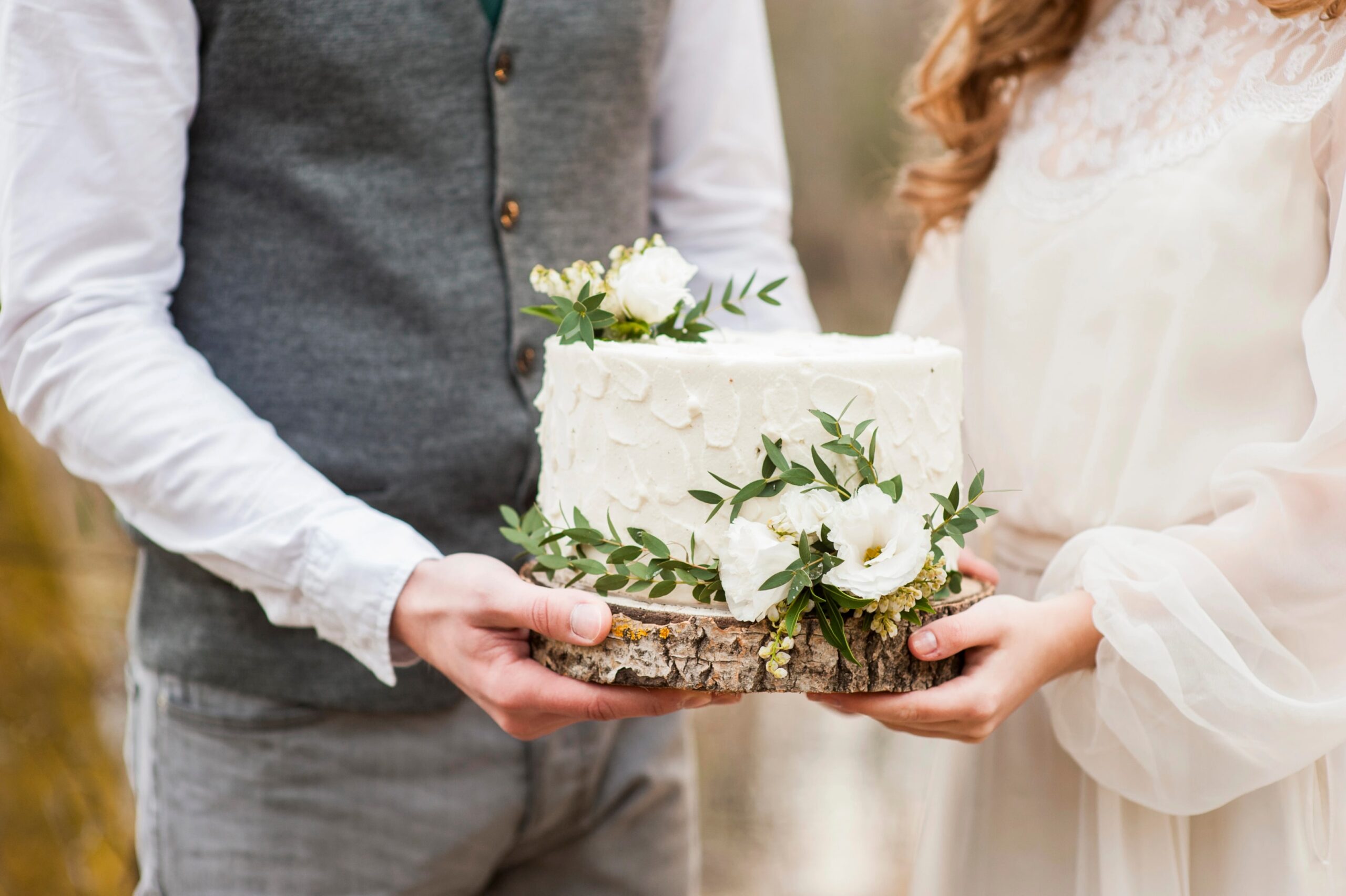 A bride and groom holding a small white wedding cake decorated with greenery and florals during a spring wedding in a park. Photo by alla-foto-alla.ru, licensed via Depositphotos.