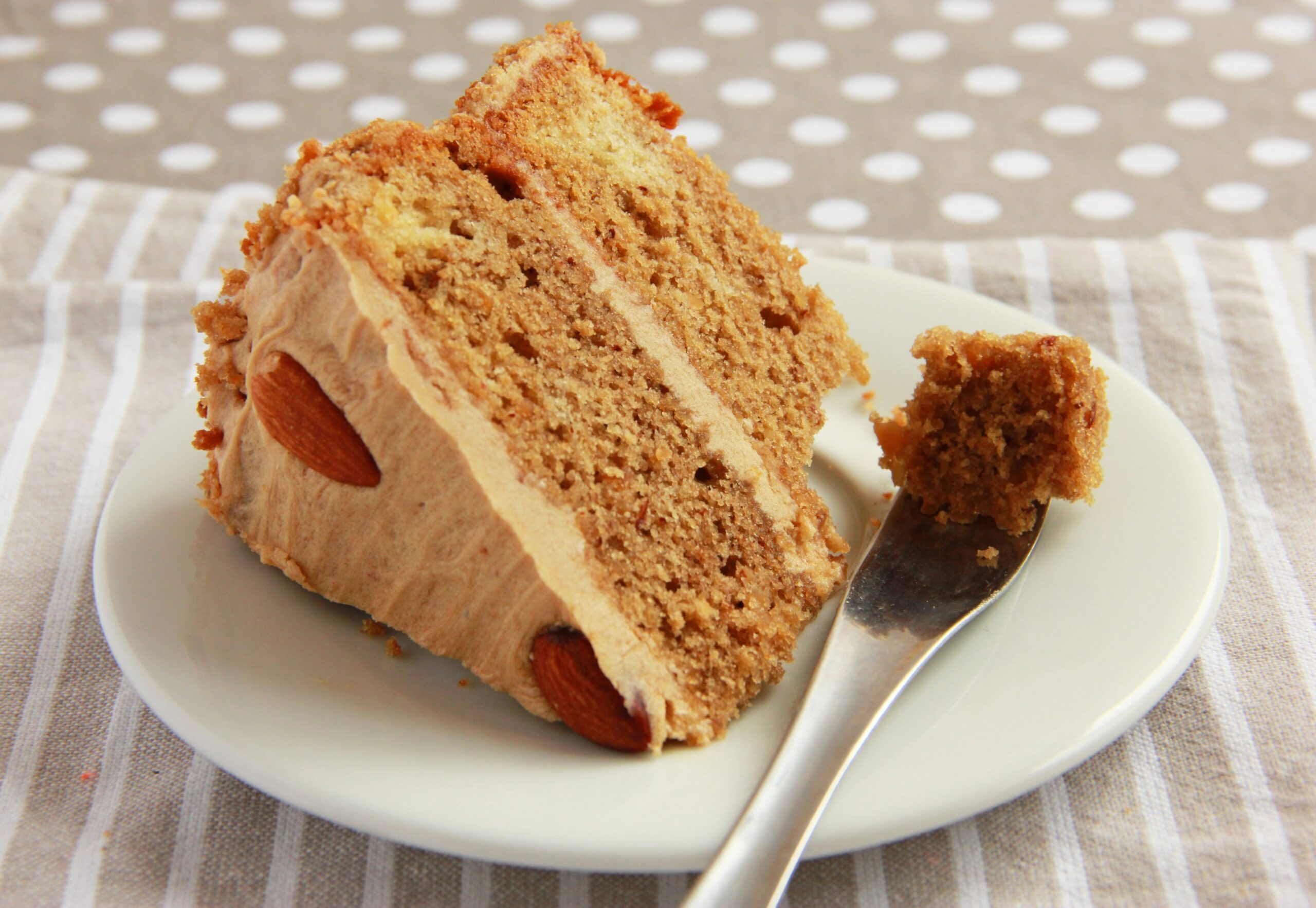 A slice of homemade almond cake layered with almond and coffee cream, served on a white plate with a fork against a soft, neutral background. Photo by chinchy, licensed via Depositphotos.