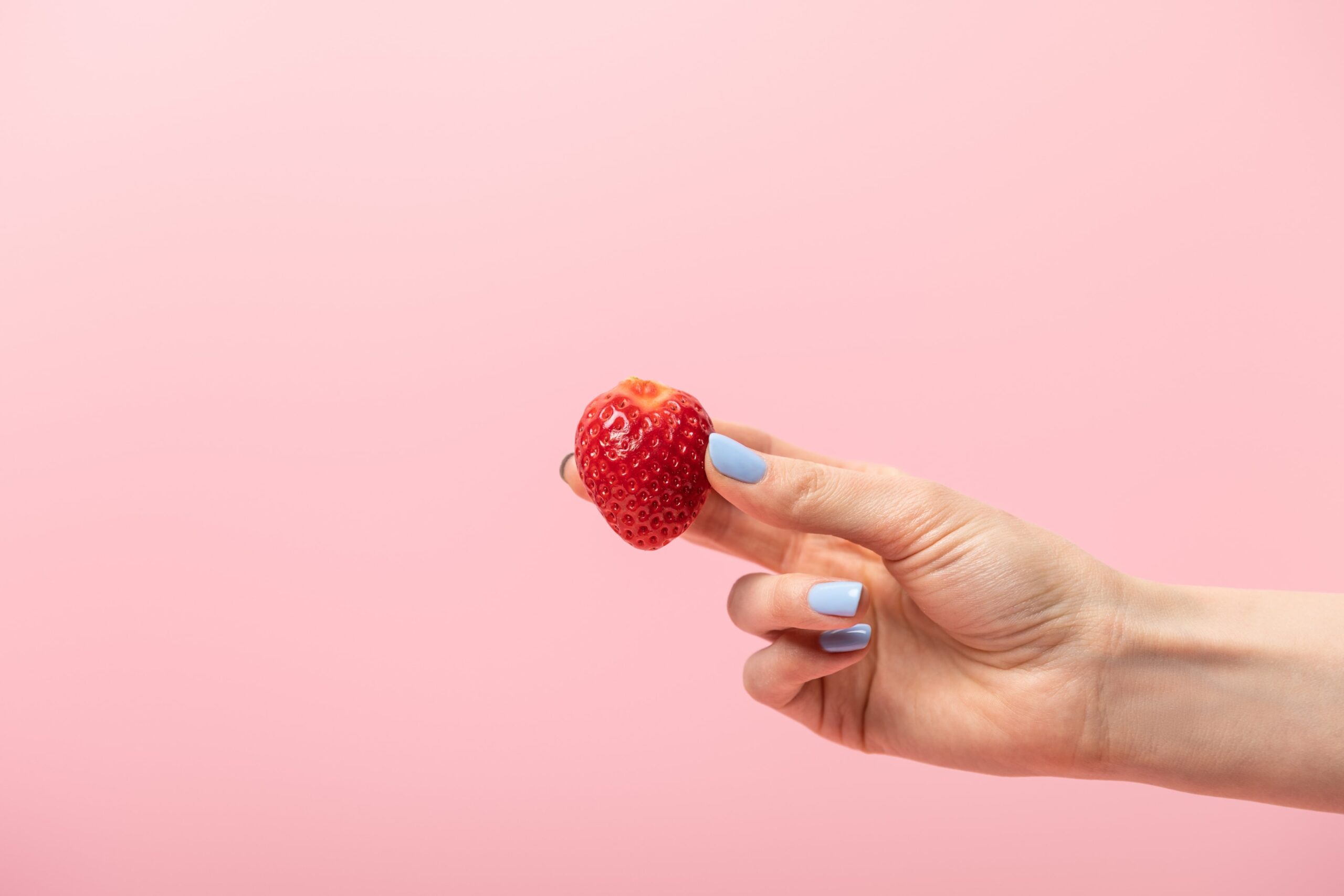 A cropped view of a hand holding a ripe red strawberry against a pastel pink background, creating a clean and colorful food detail image. Photo by AntonMatyukha, licensed via Depositphotos.