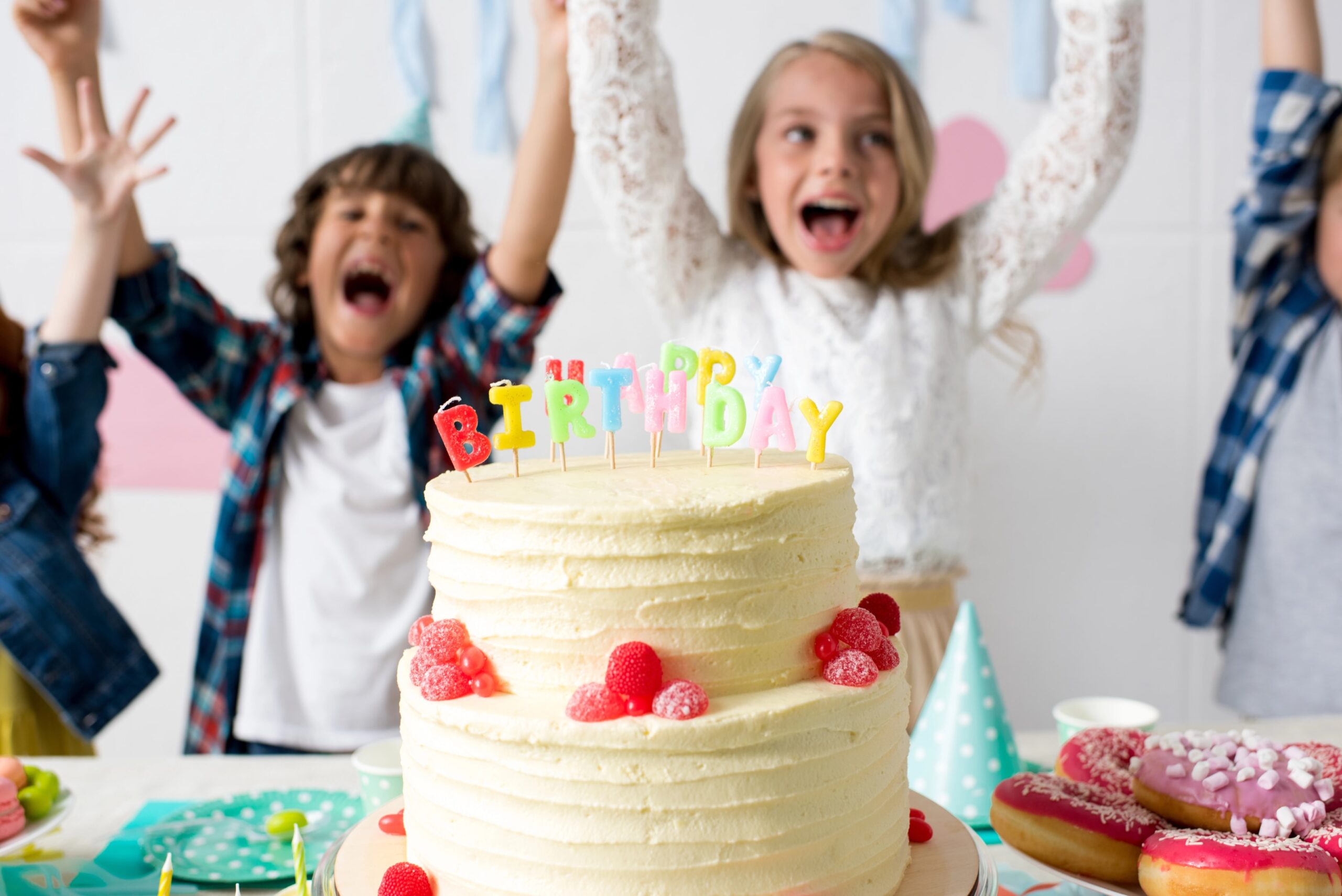 A group of happy children cheering with raised hands behind a layered birthday cake decorated with candles and fruit during a festive celebration. Photo by AlexLipa, licensed via Depositphotos.