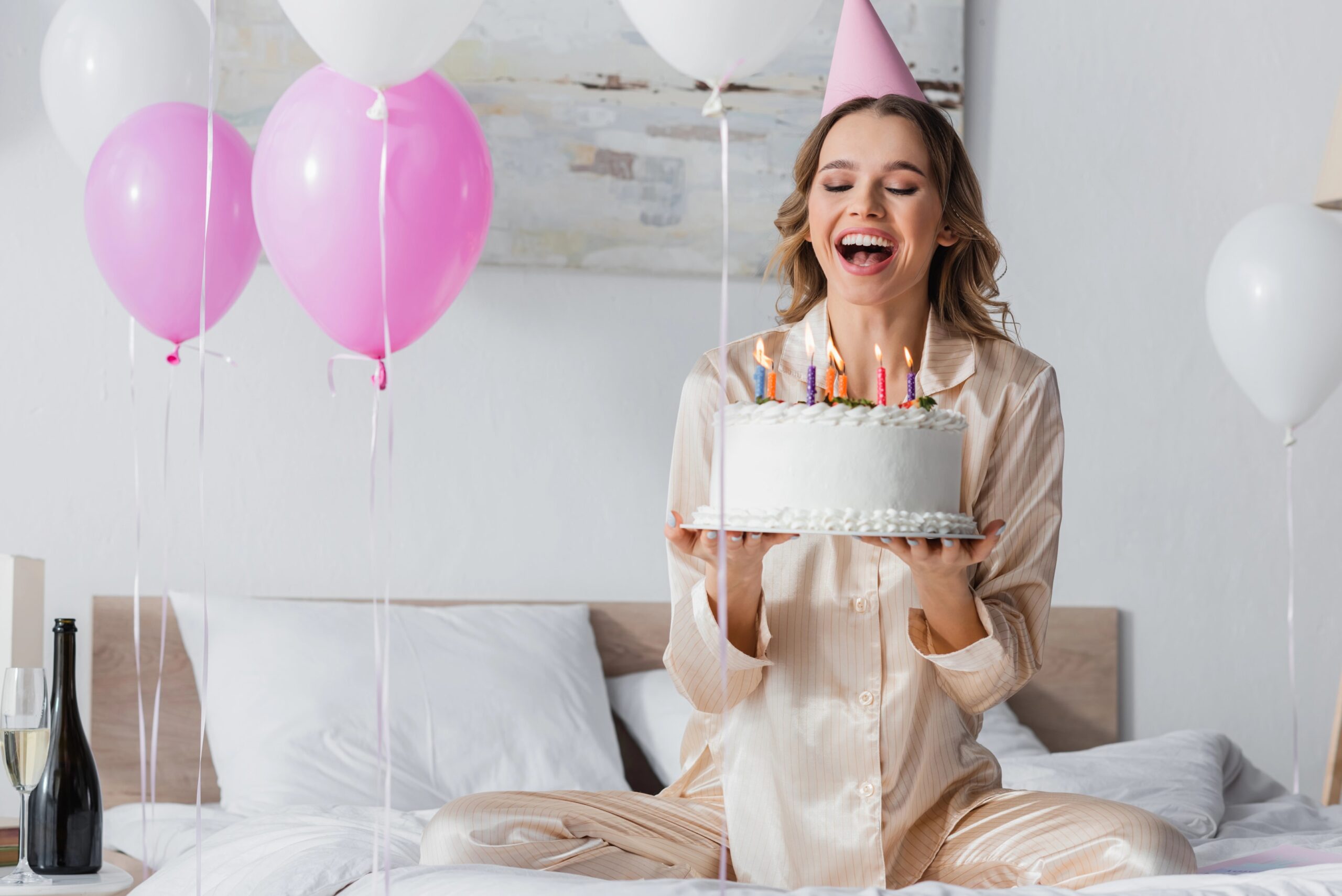 A happy woman wearing a party hat holds a birthday cake with lit candles, surrounded by pink and white balloons in a cozy indoor setting. Photo by HayDmitriy, licensed via Depositphotos.