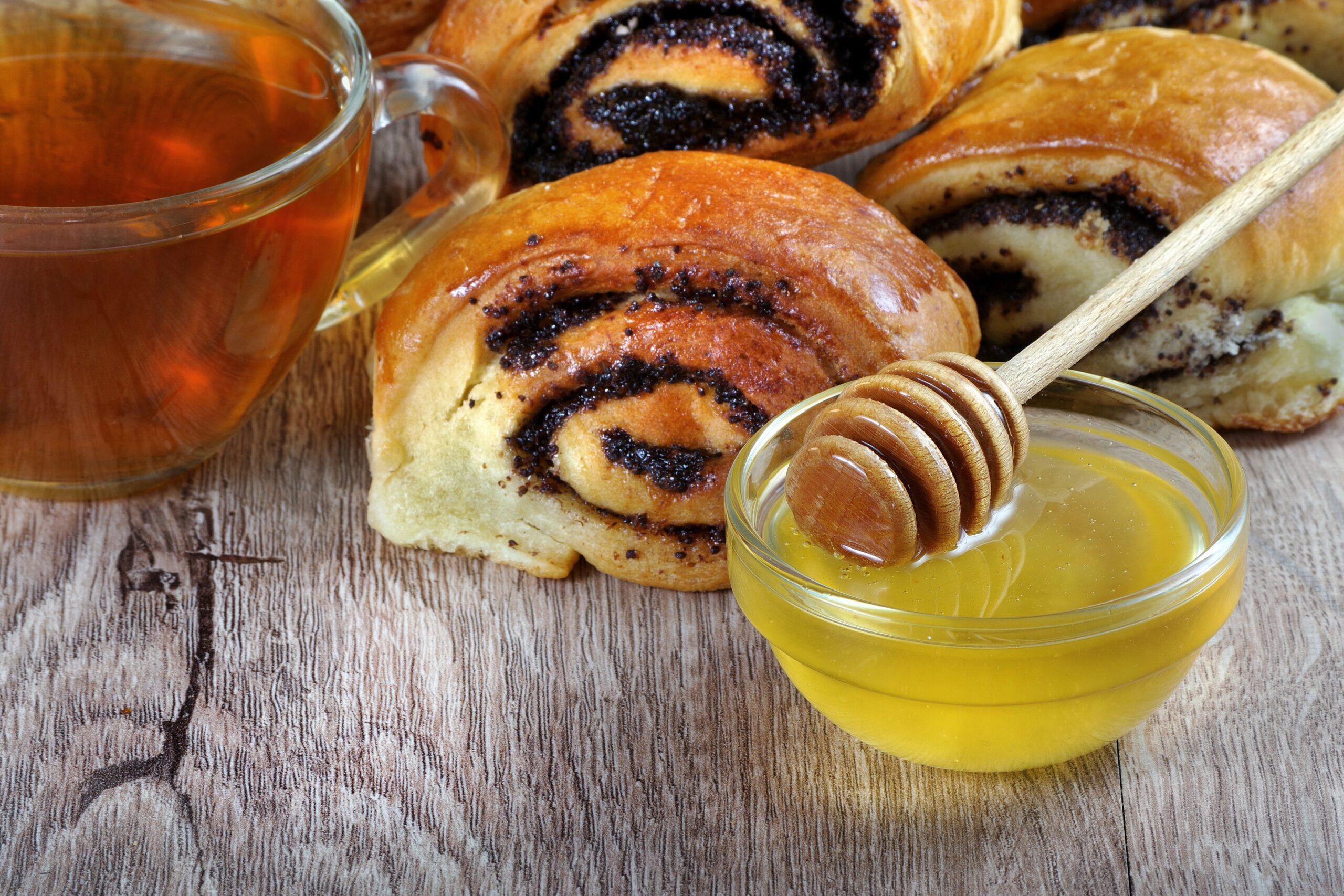 Freshly baked honey buns filled with poppy seeds, arranged on a wooden table alongside a cup of tea and a bowl of honey with a honey dipper. Photo by krjaki1973, licensed via Depositphotos.