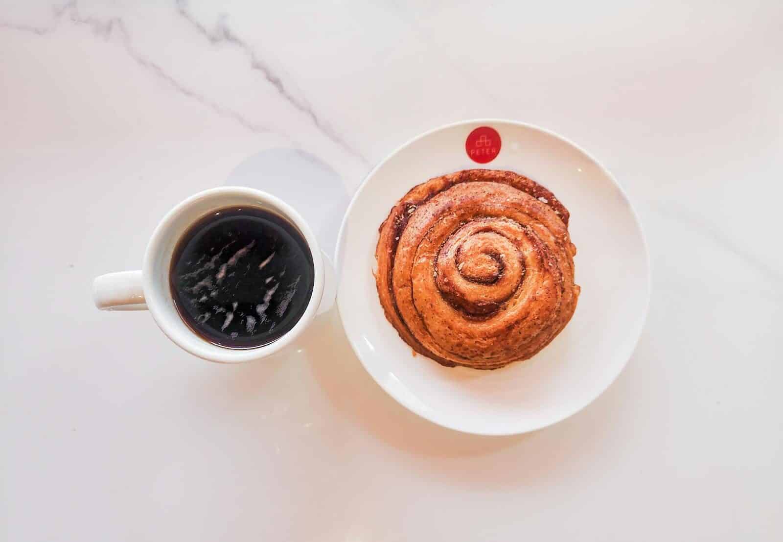 A top-view shot of black coffee with a cinnamon roll on a marble table surface.